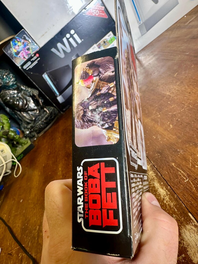 Puede incluir: Una caja de figuras de acci&oacute;n de Star Wars: El libro de Boba Fett. La caja es negra con texto rojo y blanco. El lateral de la caja presenta una imagen de un personaje de la serie. La caja est&aacute; sujeta por una persona.