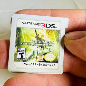 Könnte beinhalten: Eine weiße Nintendo 3DS-Spielkarte für Ace Combat Assault Horizon Legacy Plus. Das Etikett zeigt eine gelb-grüne Kampfszene, die ESRB-Einstufung Teen, das Bandai Namco-Logo und den Produktcode LNA-CTR-BCRE-USA, der unten aufgedruckt ist.