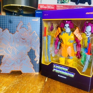 May include: A Transformers Bludgeon action figure in a Super7 box. The figure is yellow, orange, and purple with a skull-like head and comes with two swords. The box features the Transformers logo and a samurai-style illustration.