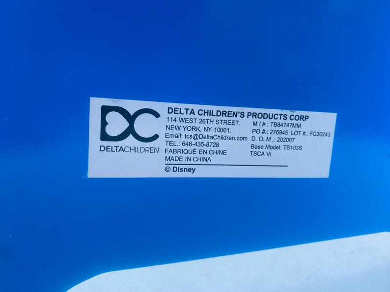 Puede incluir: Una etiqueta blanca con texto negro sobre un fondo azul. La etiqueta incluye el texto "DELTA CHILDREN'S PRODUCTS CORP", direcci&oacute;n, informaci&oacute;n de contacto y "&copy; Disney". La etiqueta tambi&eacute;n incluye el logotipo de Delta Children.