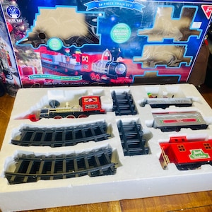 May include: A 54-piece toy train set with a red and black locomotive, train cars, and tracks. The box features a festive design with the words "North Pole Junction". The set includes a red caboose with "Merry Christmas" on it.