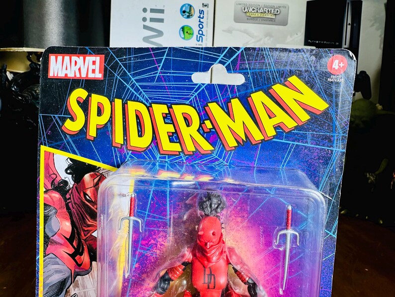 K&ouml;nnte beinhalten: Marvel Spider-Man Actionfigur in einer Kunststoffverpackung. Die Verpackung zeigt das Marvel-Logo und die Worte "Spider-Man" in Gelb und Rot. Die Figur ist eine rote Figur mit zwei silbernen Schwertern.