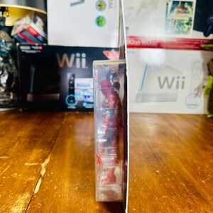 K&ouml;nnte beinhalten: Seitenansicht einer Deadpool-Actionfigur in einer transparenten Kunststoffverpackung. Die Figur ist rot und schwarz. Die Verpackung ist wei&szlig; und hat einen roten Akzent. Der Hintergrund umfasst Wii-Spielboxen und eine Holzoberfl&auml;che.