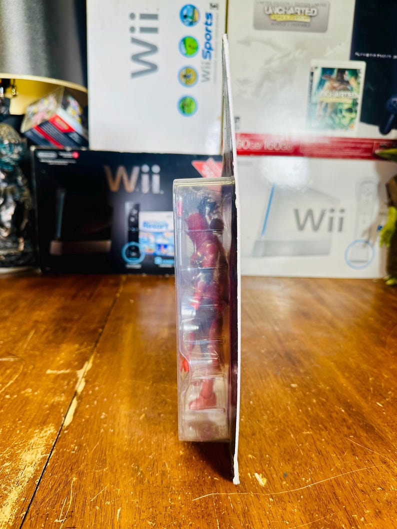 K&ouml;nnte beinhalten: Seitenansicht einer Deadpool-Actionfigur in einer transparenten Kunststoffverpackung. Die Figur ist rot und schwarz. Die Verpackung ist wei&szlig; und hat einen roten Akzent. Der Hintergrund umfasst Wii-Spielboxen und eine Holzoberfl&auml;che.