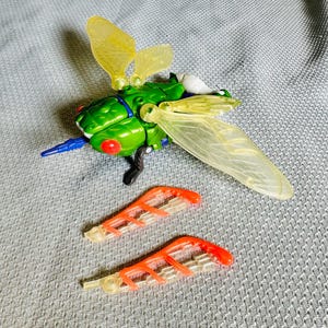 Pode incluir: Um brinquedo verde e amarelo semelhante a um inseto com asas translúcidas e acessórios de asa laranja e transparentes. O brinquedo tem olhos vermelhos e detalhes azuis e pretos. O brinquedo está sobre uma superfície cinza.