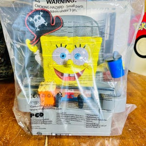 May include: A yellow SpongeBob SquarePants figure in a plastic bag. The figure is holding a blue cup and a pirate hat. The packaging includes the text "CHOKING HAZARD - Small parts. Not for children under 3 yrs."