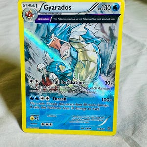 May include: A holographic Gyarados trading card with 130 HP, featuring a blue sea serpent illustration and a trainer in the foreground. The card displays the attacks Full Retaliation and Thrash, with text detailing game mechanics and card information.