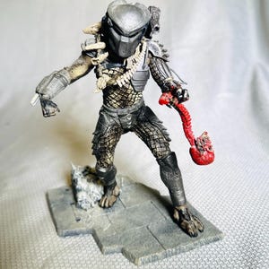Può includere: Action figure dettagliata di un alieno Predator, in piedi su una base di pietra grigia. La figura è principalmente argentata e nera, con un'armatura intricata e una maschera. Tiene in mano un oggetto rosso organico e ha ornamenti ossei.