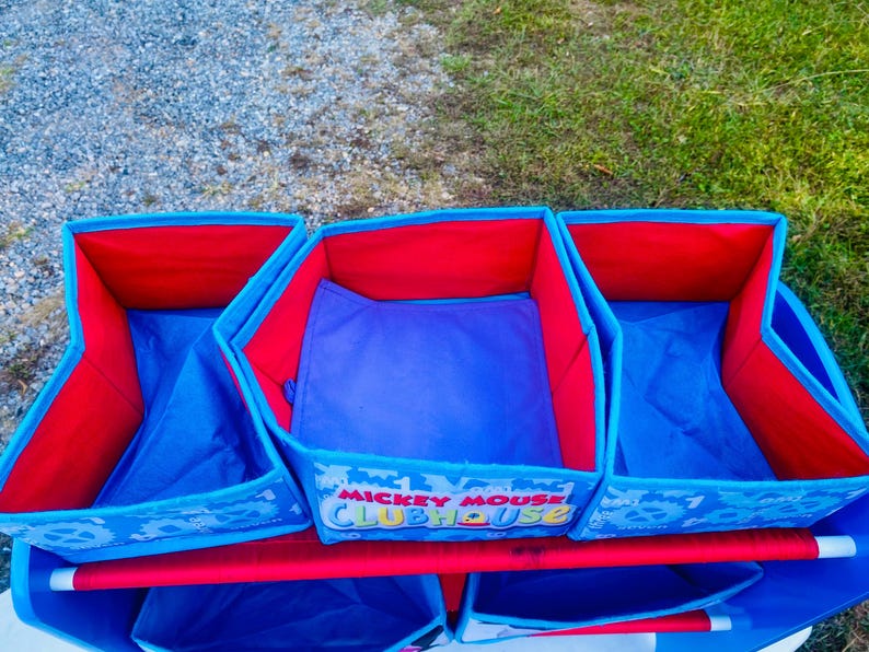 Puede incluir: Un organizador de almacenamiento de tela azul y roja con tres compartimentos. El interior de cada compartimento es rojo, con laterales azules y una base azul. El organizador tiene las palabras "Mickey Mouse Clubhouse" impresas en la parte delantera.