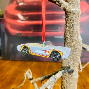 Può includere: Un'auto giocattolo cabriolet azzurra con decalcomanie a fiamma gialle e rosse, appesa a un nastro rosso. L'auto ha ruote nere con cerchi gialli. L'ornamento è appeso a un piccolo ramo.