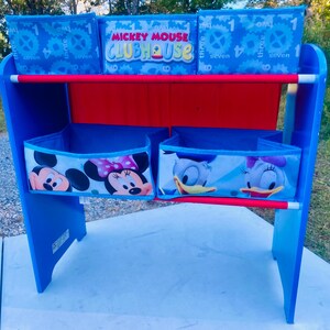 Puede incluir: Estanter&iacute;a infantil azul y roja con contenedores de tela. Los contenedores superiores tienen un dise&ntilde;o de engranajes y n&uacute;meros. Los contenedores inferiores muestran a Mickey Mouse, Minnie Mouse y Daisy Duck. Altura aproximada: 76 cm.