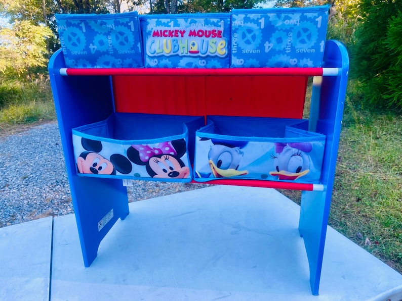 Puede incluir: Estanter&iacute;a infantil azul y roja con contenedores de tela. Los contenedores superiores tienen un dise&ntilde;o de engranajes y n&uacute;meros. Los contenedores inferiores muestran a Mickey Mouse, Minnie Mouse y Daisy Duck. Altura aproximada: 76 cm.