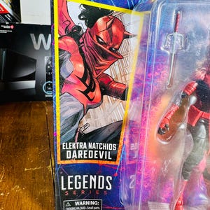 K&ouml;nnte beinhalten: Verpackte Elektra Natchios Daredevil Actionfigur aus der Legends-Serie. Die Verpackung zeigt eine Comic-Illustration von Elektra in einem rot-schwarzen Kost&uuml;m. Die Figur ist durch ein transparentes Kunststofffenster sichtbar.