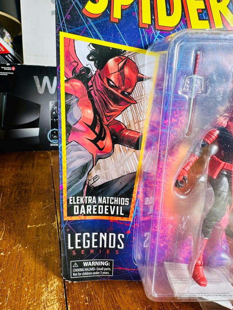 K&ouml;nnte beinhalten: Verpackte Elektra Natchios Daredevil Actionfigur aus der Legends-Serie. Die Verpackung zeigt eine Comic-Illustration von Elektra in einem rot-schwarzen Kost&uuml;m. Die Figur ist durch ein transparentes Kunststofffenster sichtbar.
