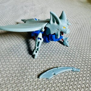 Puede incluir: Una figura de acción de plástico con forma de murciélago en color gris y azul, con orejas puntiagudas y ojos amarillos. La figura presenta alas y extremidades articuladas, con un componente curvo de plástico gris separado frente al cuerpo.