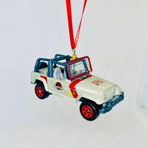 May include: A miniature off-white Jeep Wrangler ornament with red and blue accents, featuring the text "JURASSIC PARK". The ornament has red wheels and a red ribbon for hanging. A fun collectible for fans of the movie.