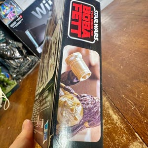 Puede incluir: Una caja negra con el logotipo de Star Wars: The Book of Boba Fett en rojo y blanco. La caja muestra una imagen de un objeto dorado y una figura p&uacute;rpura. La caja est&aacute; sostenida por una persona.