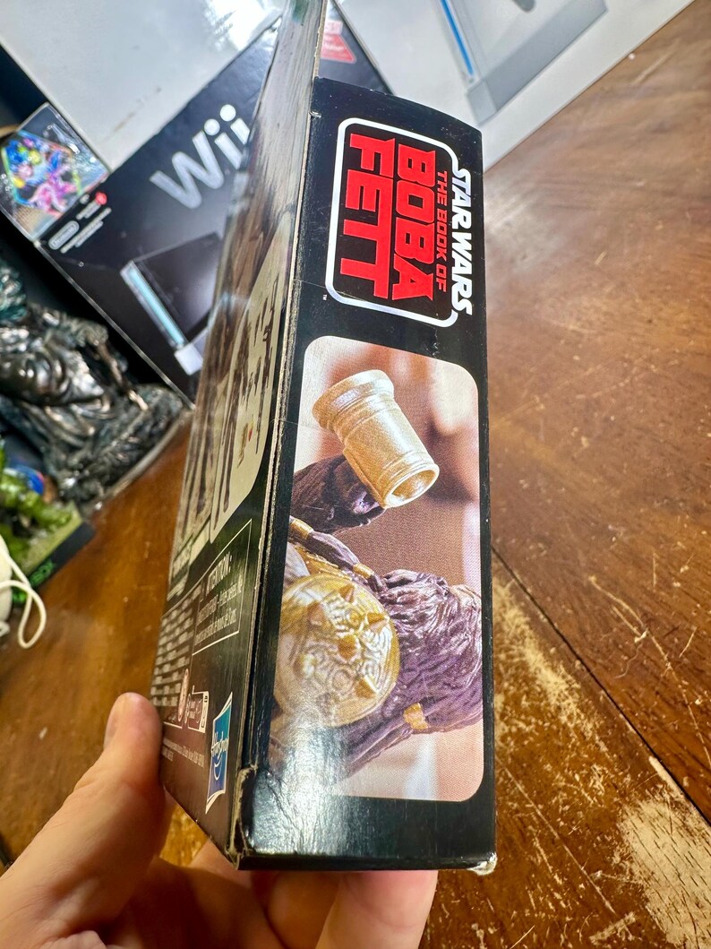 Puede incluir: Una caja negra con el logotipo de Star Wars: The Book of Boba Fett en rojo y blanco. La caja muestra una imagen de un objeto dorado y una figura p&uacute;rpura. La caja est&aacute; sostenida por una persona.