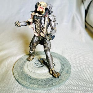 May include: A detailed Predator action figure, standing on a circular base. The figure is predominantly silver and brown, with intricate armor and a detailed face. The base has a textured, gray design.