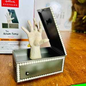 Può includere: Un ornamento Hallmark Keepsake di "Mano" della Famiglia Addams. La mano esce da una scatola nera con coperchio incernierato, rifinita con perle finte bianche. La scatola è grigia con un pomello nero.