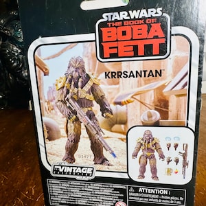 Puede incluir: Figura de acci&oacute;n de Star Wars El Libro de Boba Fett Krrsantan. La figura es marr&oacute;n y beige con detalles dorados y un rifle bl&aacute;ster. El embalaje incluye el logotipo de Vintage Collection y el logotipo de Disney.
