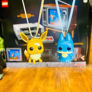 Puede incluir: Dos collares con personajes de Pokemon. El collar de la izquierda presenta un Eevee marrón con una cola amarilla. El collar de la derecha presenta un Gengar azul con un ojo amarillo.