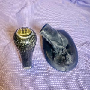 May include: A black carbon fibre gear shift knob with a gold gear shift pattern and a black leather gear shift boot.