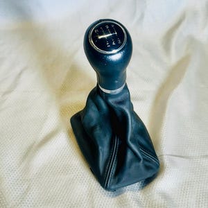 May include: A black gear stick knob with a chrome ring and a leather gaiter. The knob displays a gear position diagram. The gaiter is made of black leather and is stitched.