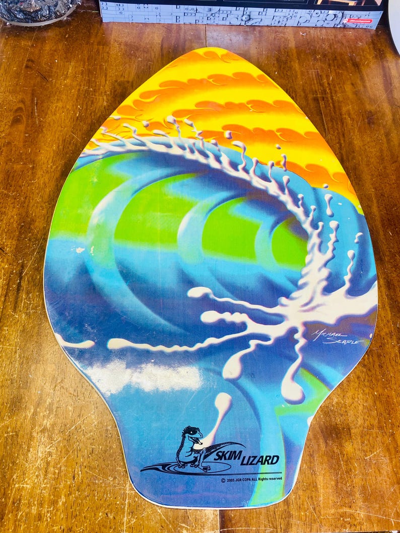 May include: A blue, green, and yellow skimboard with a wave design and a white splash of water. The board has a black lizard logo with the text "Skim Lizard" and the copyright information "2003 Jon COPE All rights reserved".