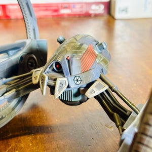 May include: A detailed model of a Separatist Droid Tri-fighter from the Star Wars universe. The model features a gray and blue color scheme with red accents and a red eye. The model has multiple legs and a central body with the Separatist symbol.