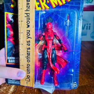 K&ouml;nnte beinhalten: Marvel Elektra Daredevil Actionfigur in einer Kunststoffverpackung. Die Figur ist rot und schwarz, mit einer roten Maske und einem Schwert-Zubeh&ouml;r. Die Verpackung zeigt das Spider-Man-Logo und den Text "Do unto others as you would have them."
