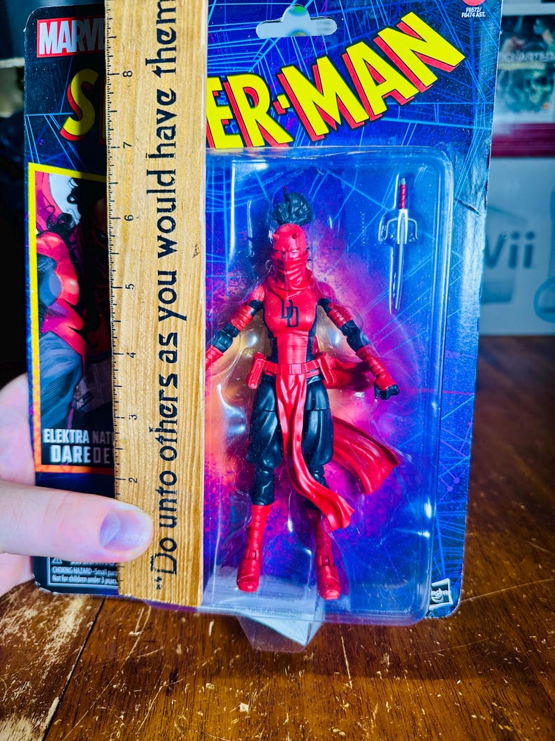 K&ouml;nnte beinhalten: Marvel Elektra Daredevil Actionfigur in einer Kunststoffverpackung. Die Figur ist rot und schwarz, mit einer roten Maske und einem Schwert-Zubeh&ouml;r. Die Verpackung zeigt das Spider-Man-Logo und den Text "Do unto others as you would have them."