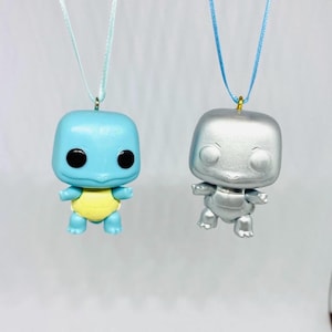 Puede incluir: Dos pequeños adornos de figuritas, uno azul y otro plateado, colgando de una cinta azul. Las figuritas tienen forma de tortuga con el vientre amarillo.