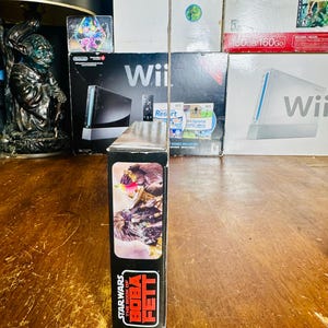 Puede incluir: Una caja negra para un producto de Star Wars: The Book of Boba Fett. La caja presenta una imagen de Boba Fett y el logotipo de Star Wars. La caja se muestra frente a varias cajas de juegos de Wii.