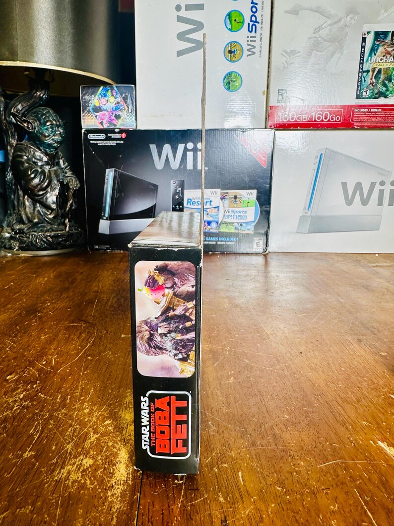 Puede incluir: Una caja negra para un producto de Star Wars: The Book of Boba Fett. La caja presenta una imagen de Boba Fett y el logotipo de Star Wars. La caja se muestra frente a varias cajas de juegos de Wii.