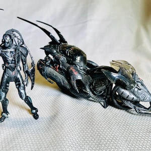 Puede incluir: Figura de acción gris metálico de un alienígena humanoide con múltiples apéndices, junto a una motocicleta futurista. La figura tiene un diseño detallado con casco y armadura. La motocicleta es negra con parabrisas transparente.