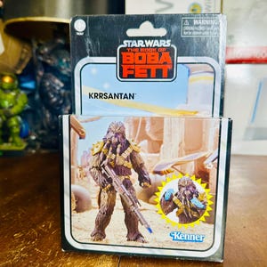 Puede incluir: Figura de acci&oacute;n de Star Wars: The Book of Boba Fett de Krrsantan en su embalaje original. La figura es marr&oacute;n con detalles dorados y sostiene un bl&aacute;ster. El embalaje presenta el logotipo de Star Wars y la marca Kenner.