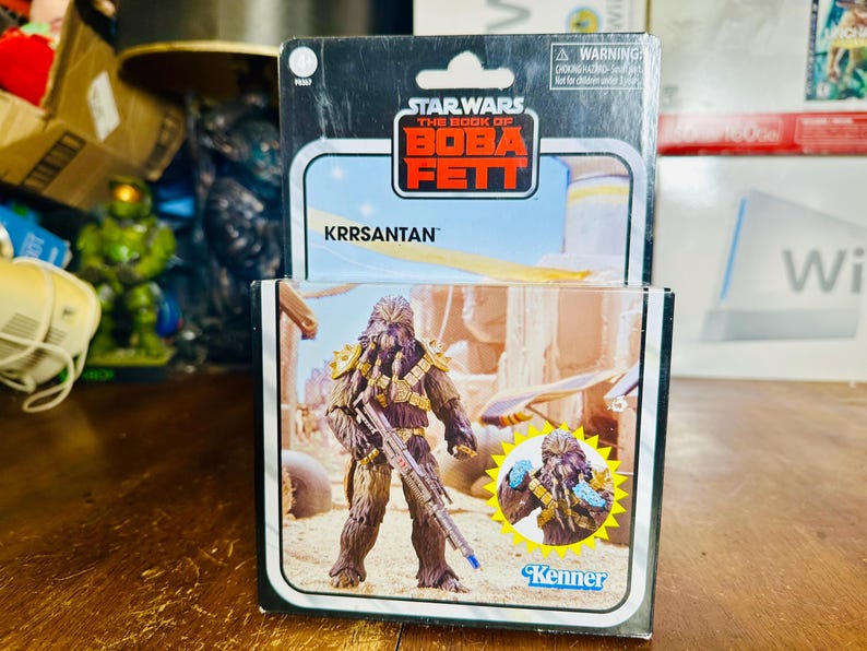 Puede incluir: Figura de acci&oacute;n de Star Wars: The Book of Boba Fett de Krrsantan en su embalaje original. La figura es marr&oacute;n con detalles dorados y sostiene un bl&aacute;ster. El embalaje presenta el logotipo de Star Wars y la marca Kenner.