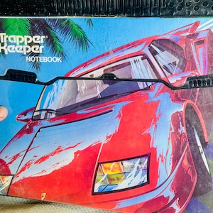 May include: A Trapper Keeper notebook with a vibrant red sports car design. The notebook cover features the text "Trapper Keeper NOTEBOOK" and a tropical palm tree illustration against a blue background.
