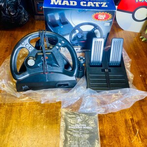 May include: A black Mad Catz analog steering wheel set for Nintendo 64, including a steering wheel, pedals, and a user's guide. The box features the Mad Catz logo and product details. The set is new and in its original packaging.