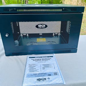 Puede incluir: Un armario SmartRack Tripp Lite negro con un panel frontal de malla y una cerradura. El armario está sobre una superficie blanca, con un manual del propietario delante. El manual se titula "Non-Swinging Wall-Mounted SmartRack Enclosures."