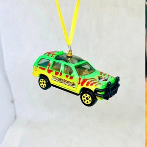 May include: A green and yellow toy SUV with the text "JURASSIC PARK" and red paint splatters. The toy car has black wheels and a black front bumper. A yellow ribbon is attached to the top of the car.