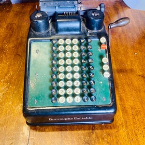 Può includere: Un'antica macchina da calcolo Burroughs Portable. La macchina nera ha una tastiera verde con tasti numerici bianchi e neri. Il testo "Burroughs Portable" è stampato sul davanti.