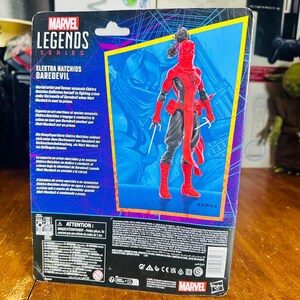 K&ouml;nnte beinhalten: Marvel Legends Series Actionfigur von Elektra Natchios Daredevil, in einem rot-schwarzen Kost&uuml;m mit Sai-Waffen. Die Figur ist in einer blau-violetten Box mit Charakterdetails und dem Marvel-Logo verpackt.