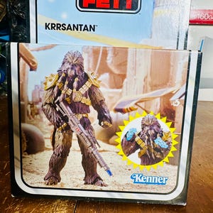 Puede incluir: Una caja de figuras de acci&oacute;n Kenner vintage con el personaje Krrsantan de Star Wars. El arte de la caja representa al guerrero Wookiee sosteniendo un rifle bl&aacute;ster, ambientado en un desierto. La figura es marr&oacute;n con detalles dorados. El logotipo de Kenner es visible.