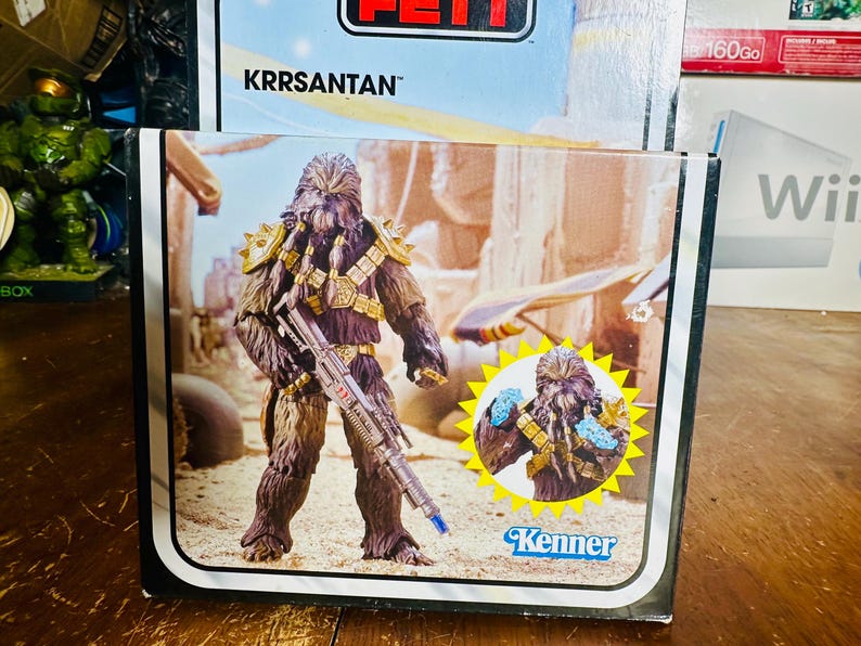 Puede incluir: Una caja de figuras de acci&oacute;n Kenner vintage con el personaje Krrsantan de Star Wars. El arte de la caja representa al guerrero Wookiee sosteniendo un rifle bl&aacute;ster, ambientado en un desierto. La figura es marr&oacute;n con detalles dorados. El logotipo de Kenner es visible.