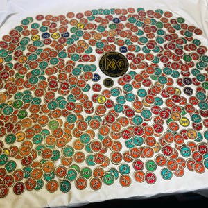 Può includere: Una collezione di toppe rotonde colorate con la scritta "POG" e numeri, in tonalità di rosso, arancione, verde e blu. È presente anche una toppa più grande con "POG" e "DOM AGAINST FOR TERROR". Le toppe sono su una superficie bianca.