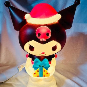 Puede incluir: Figura iluminada de un personaje de dibujos animados con cabeza negra, orejas grandes y gorro de Papá Noel rojo. El personaje sostiene un regalo con un lazo azul. La figura tiene una base blanca y está iluminada desde dentro.