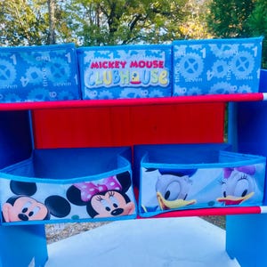 Puede incluir: Una estanter&iacute;a azul y roja de Mickey Mouse Clubhouse con contenedores de almacenamiento. Los contenedores superiores tienen un fondo azul con engranajes blancos y los n&uacute;meros 1, 3, 4, 7 y 8. El contenedor central tiene las palabras "Mickey Mouse Clubhouse". Los contenedores inferiores presentan a Mickey, Minnie, Donald y Daisy.