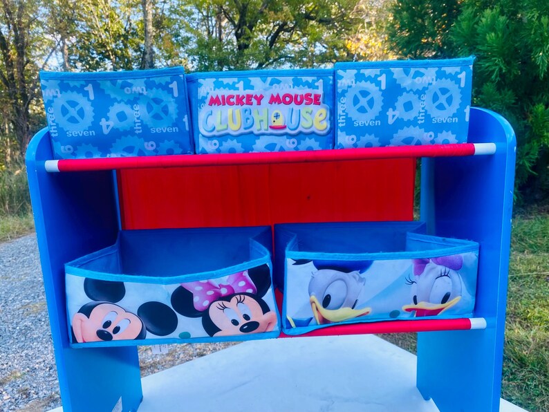 Puede incluir: Una estanter&iacute;a azul y roja de Mickey Mouse Clubhouse con contenedores de almacenamiento. Los contenedores superiores tienen un fondo azul con engranajes blancos y los n&uacute;meros 1, 3, 4, 7 y 8. El contenedor central tiene las palabras "Mickey Mouse Clubhouse". Los contenedores inferiores presentan a Mickey, Minnie, Donald y Daisy.